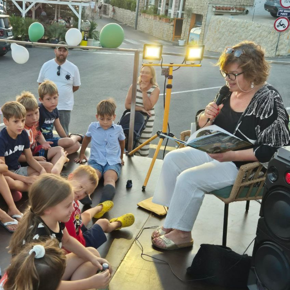 Ljeto u Metajni 2025: Maja Cimbal čita djeci na rivi Metajne priču iz „Priče iz Novalje – David otkriva legendu o Caskoj“, uz pjesmu i crtanje.